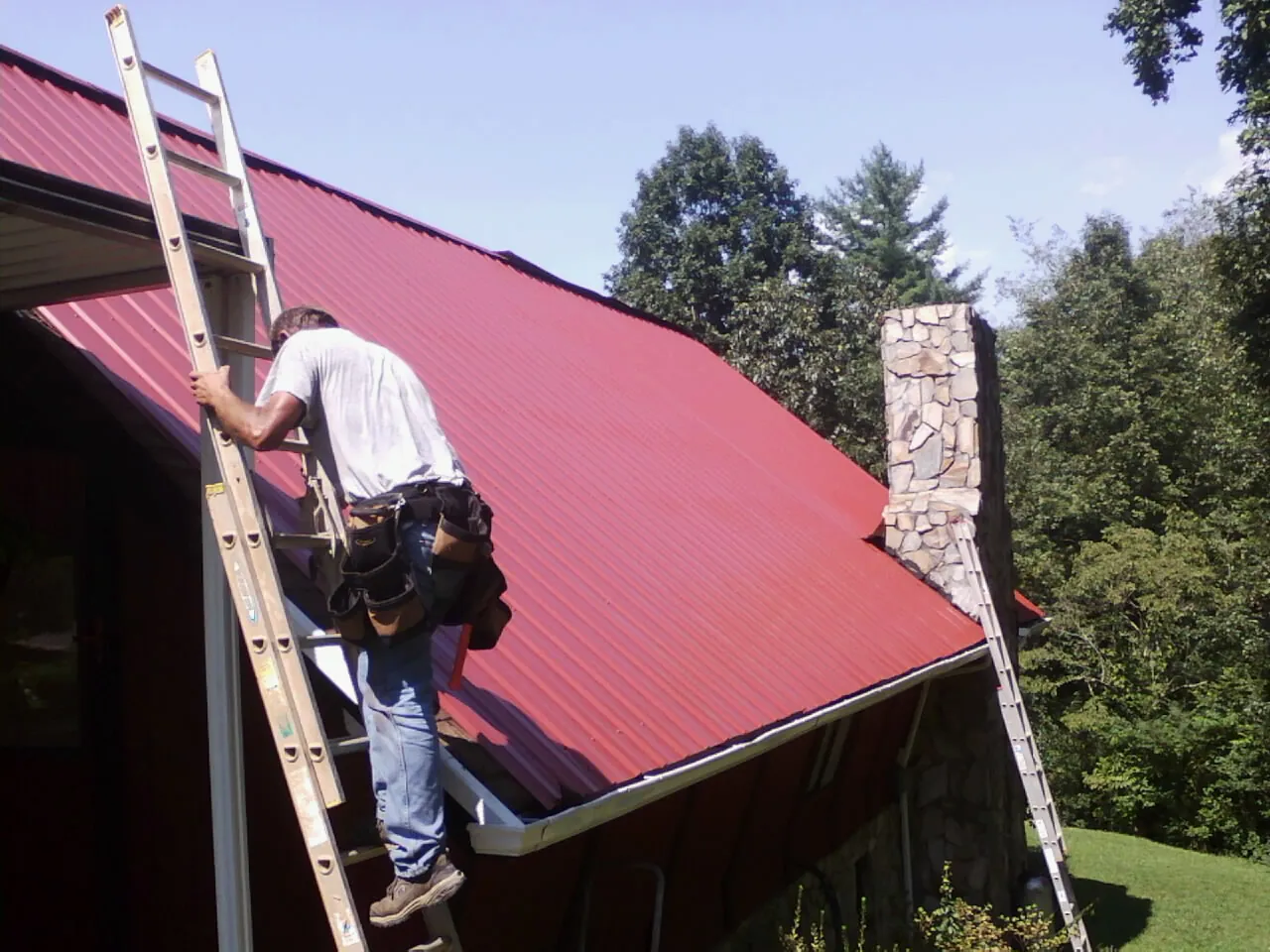 Roofer on a ladder beside a red metal roof and stone chimney — Soffit & Fascia Repair by Roofing Brothers in Jackpot